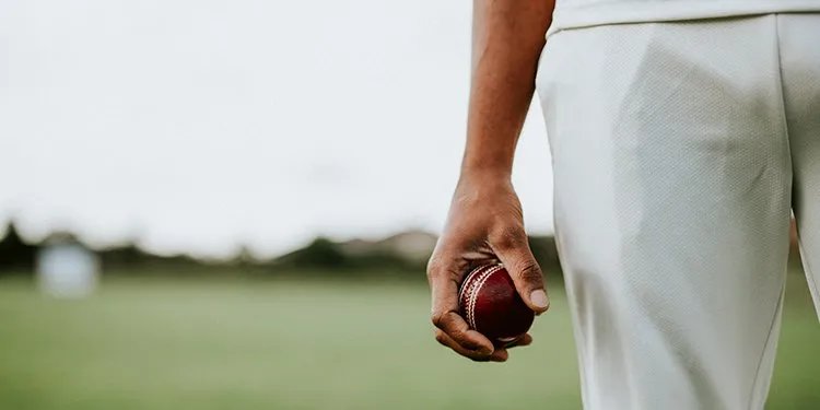 Bowler holding a cricket ball behind his back while walking, whites and grass ground visible